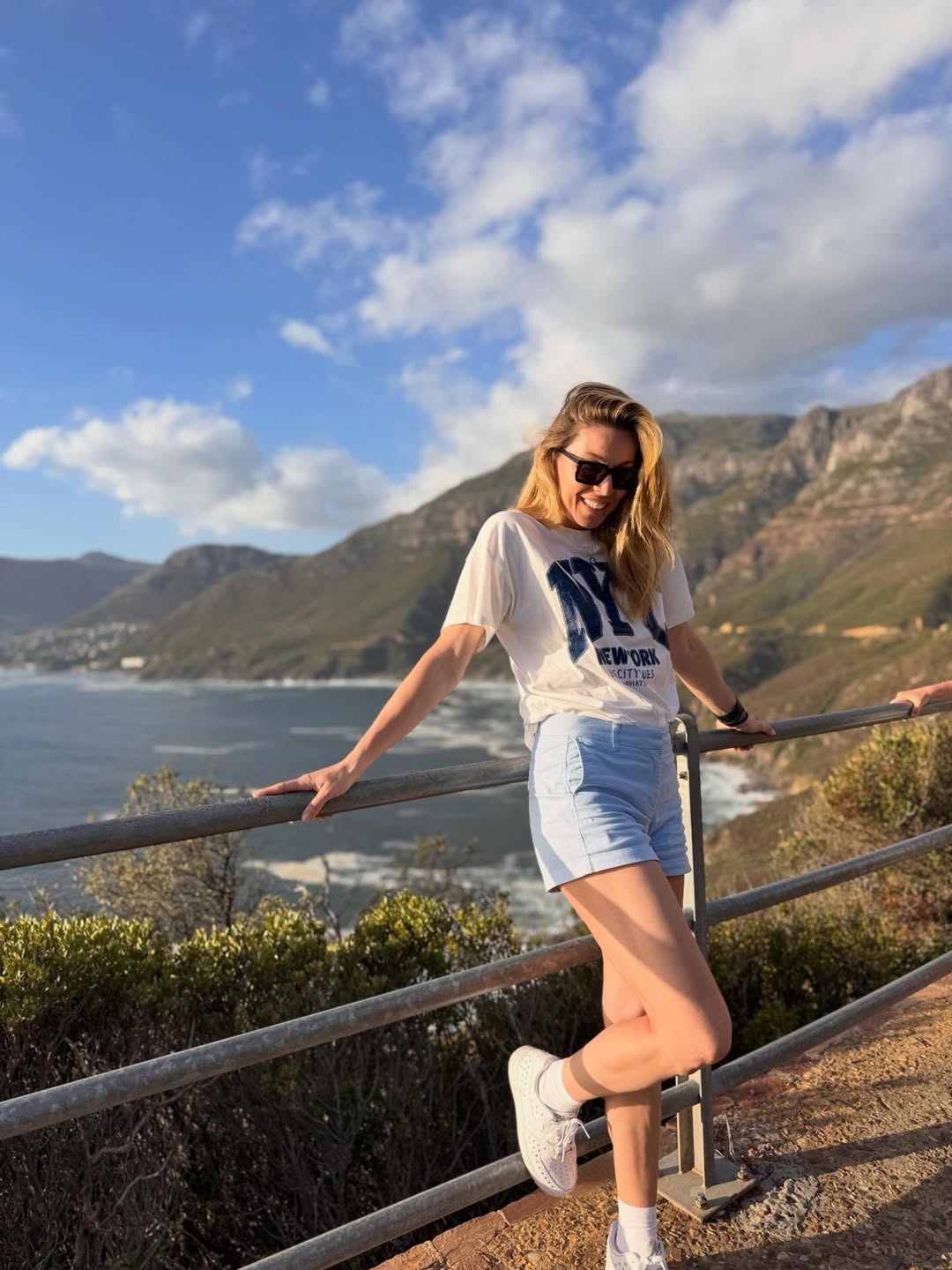 Το σκηνικό που ξεδιπλώνεται πίσω της θυμίζει καρτ ποστάλ, με εντυπωσιακές ακτές, καθαρό ορίζοντα και επιβλητικά βουνά να συνθέτουν ένα ιδανικό ταξιδιωτικό φόντο.